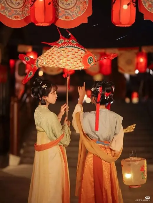 Two women in Tang-style dress admiring a fish lantern at night